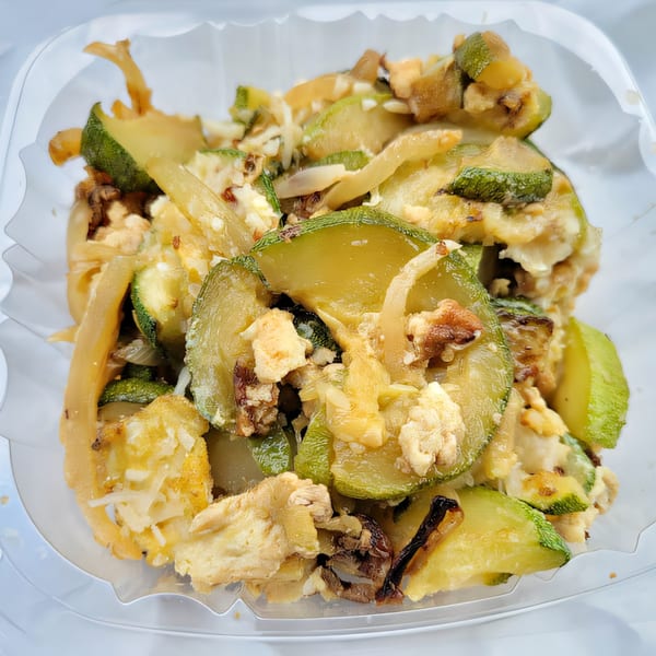 Close-up of sautéed zucchini, onions, and scrambled eggs in a clear plastic container.