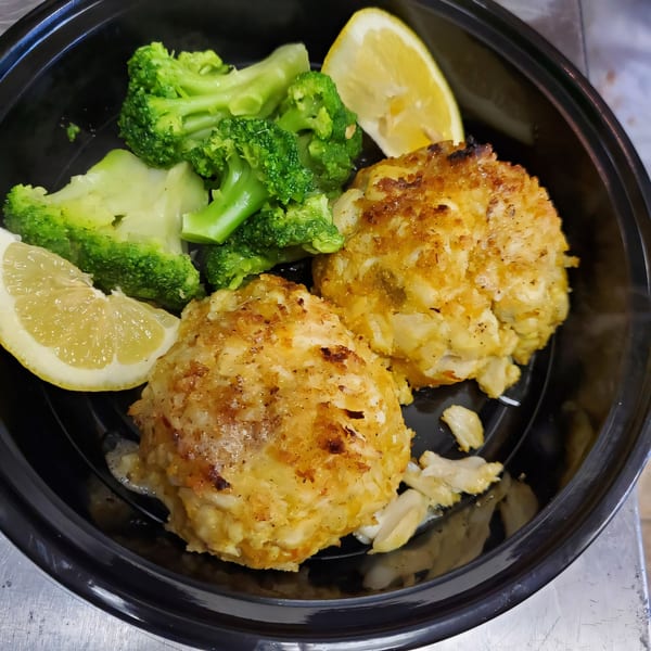 Two golden-brown crab cakes with steamed broccoli and lemon wedges in a black takeout container.