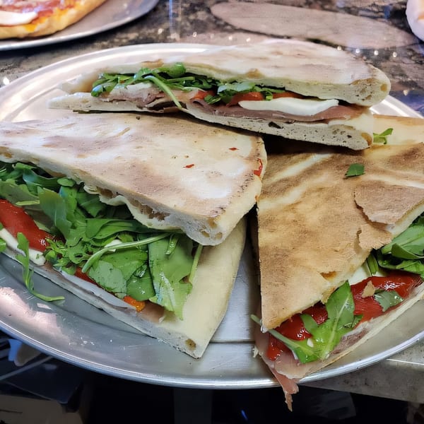 Three Italian flatbread sandwiches with prosciutto, mozzarella, roasted red peppers, and arugula on a metal tray.